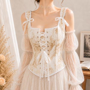 May include: A white corset top featuring a gold floral pattern, a front lace-up closure with white ribbons, and shoulder straps tied with bows. The garment includes sheer, long, puffed sleeves made of lace and a matching lace choker necklace.