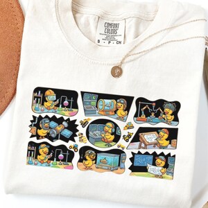 May include: Ivory-colored t-shirt featuring a graphic of a cartoon duck in various science and engineering scenarios. The shirt has a crew neck and short sleeves. A gold necklace with a pendant is draped across the shirt.