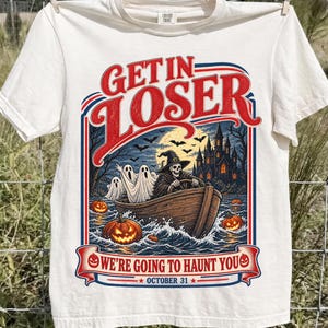 May include: An off-white short-sleeve t-shirt featuring a graphic print of a skeleton in a witch hat driving a wooden boat with ghosts. The text reads GET IN LOSER WE'RE GOING TO HAUNT YOU OCTOBER 31. The design includes pumpkins and a castle.