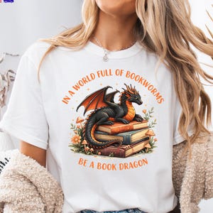 May include: A white t-shirt featuring a graphic of a black dragon with orange wings resting on a stack of books. The text reads, "IN A WORLD FULL OF BOOKWORMS BE A BOOK DRAGON," surrounded by small orange flowers and mushrooms in a circular arrangement.