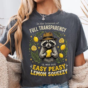 May include: A charcoal gray t-shirt featuring a graphic of a raccoon wearing a brown fedora hat and holding a yellow lemon. The text reads In the interest of FULL TRANSPARENCY it was not EASY PEASY LEMON SQUEEZY, surrounded by lemon illustrations.