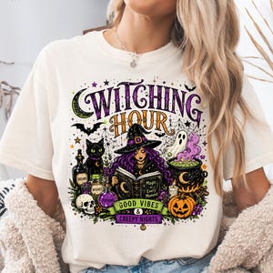May include: An ivory t-shirt featuring a graphic print with the text Witching Hour, Good Vibes & Creepy Nights. The design includes a witch reading a book, a black cat, a skull, a cauldron with purple smoke, a ghost, bats, and stars.