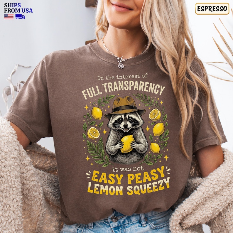 May include: A brown short-sleeve t-shirt featuring a graphic of a raccoon wearing a fedora hat and holding a lemon. The text reads In the interest of FULL TRANSPARENCY it was not EASY PEASY LEMON SQUEEZY, surrounded by yellow lemons and green leaves.