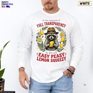 May include: A white long-sleeve shirt featuring a graphic of a raccoon wearing a brown fedora and holding a yellow lemon. The text reads: In the interest of FULL TRANSPARENCY it was not EASY PEASY LEMON SQUEEZY. Green leaves and yellow lemons surround the raccoon.