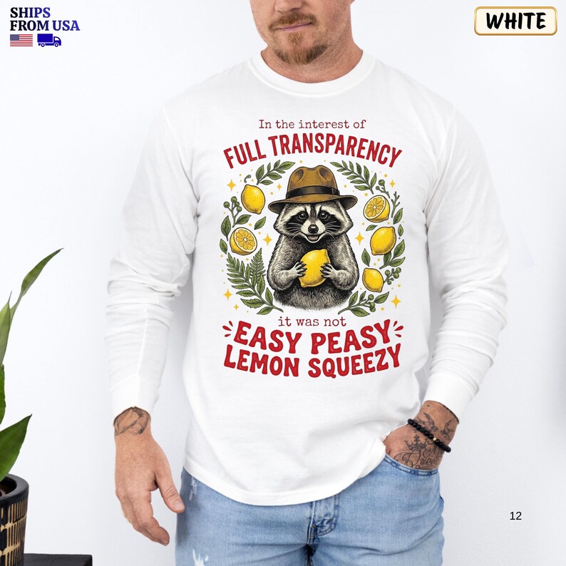 May include: A white long-sleeve shirt featuring a graphic of a raccoon wearing a brown fedora and holding a yellow lemon. The text reads: In the interest of FULL TRANSPARENCY it was not EASY PEASY LEMON SQUEEZY. Green leaves and yellow lemons surround the raccoon.