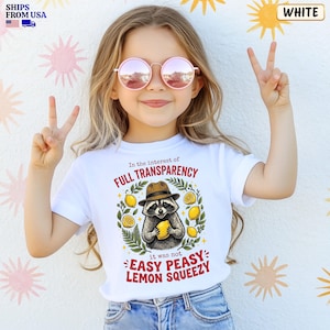 May include: A white t-shirt featuring a graphic of a raccoon wearing a brown fedora and holding a yellow lemon. The text reads In the interest of FULL TRANSPARENCY it was not EASY PEASY LEMON SQUEEZY, surrounded by green leaves and yellow lemons.