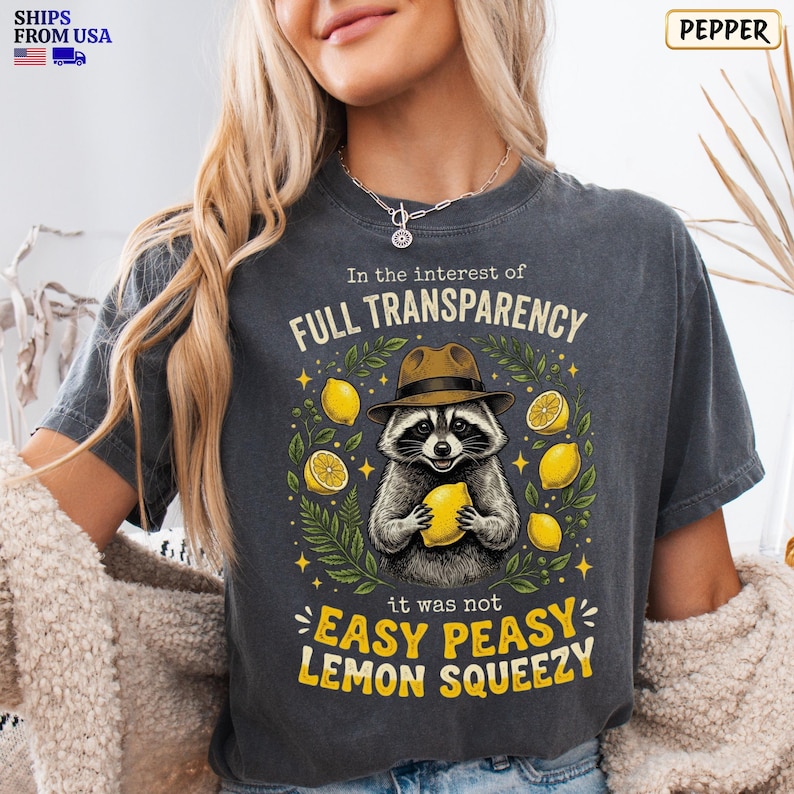 May include: A charcoal gray t-shirt featuring a graphic of a raccoon wearing a brown fedora hat and holding a yellow lemon. The text reads In the interest of FULL TRANSPARENCY it was not EASY PEASY LEMON SQUEEZY, surrounded by lemon illustrations.