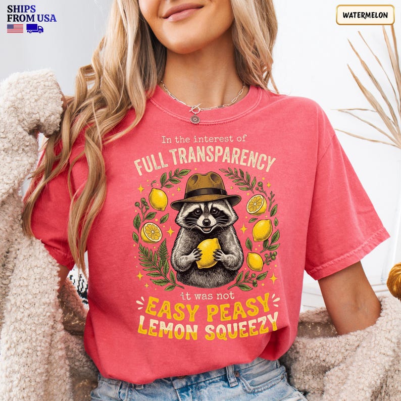 May include: A watermelon-colored t-shirt featuring a graphic of a raccoon wearing a brown hat and holding a yellow lemon. The text reads: In the interest of FULL TRANSPARENCY it was not EASY PEASY LEMON SQUEEZY. Green leaves and yellow lemons surround the raccoon.