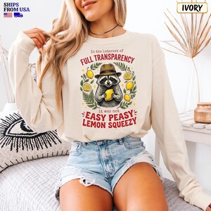 May include: An ivory long-sleeve sweatshirt featuring a graphic of a raccoon wearing a hat and holding a lemon. The text reads: In the interest of FULL TRANSPARENCY it was not EASY PEASY LEMON SQUEEZY. The design includes green leaves and yellow lemons.