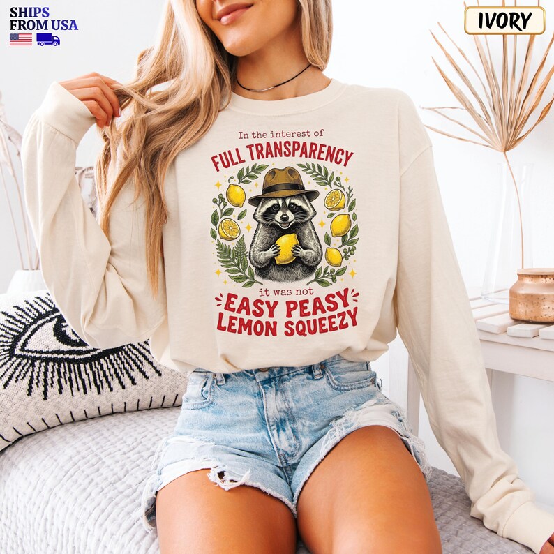 May include: An ivory long-sleeve sweatshirt featuring a graphic of a raccoon wearing a hat and holding a lemon. The text reads: In the interest of FULL TRANSPARENCY it was not EASY PEASY LEMON SQUEEZY. The design includes green leaves and yellow lemons.
