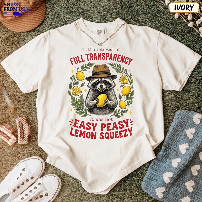 May include: An ivory short-sleeve t-shirt featuring a graphic of a raccoon wearing a brown fedora and holding a yellow lemon. The text reads In the interest of FULL TRANSPARENCY it was not EASY PEASY LEMON SQUEEZY surrounded by green leaves.