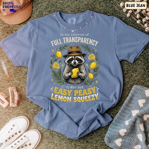 May include: A blue short-sleeve t-shirt featuring a graphic of a raccoon wearing a brown hat and holding a yellow lemon. The text reads In the interest of FULL TRANSPARENCY it was not EASY PEASY LEMON SQUEEZY, surrounded by lemon and leaf illustrations.