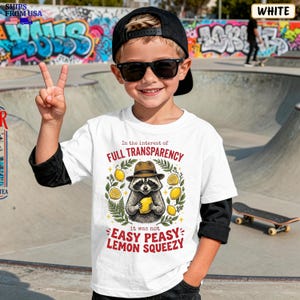 May include: A white short-sleeved t-shirt featuring a graphic of a raccoon wearing a brown fedora and holding a yellow lemon. The text reads In the interest of FULL TRANSPARENCY it was not EASY PEASY LEMON SQUEEZY, surrounded by green leaves.