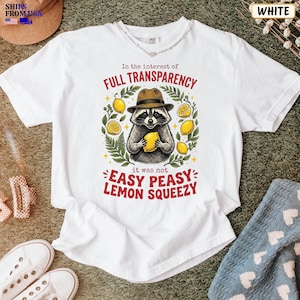 May include: A white short-sleeve t-shirt featuring a graphic of a raccoon wearing a brown hat and holding a yellow lemon. The text reads In the interest of FULL TRANSPARENCY it was not EASY PEASY LEMON SQUEEZY, surrounded by green leaves.