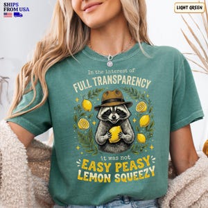 May include: A light green t-shirt featuring a graphic of a raccoon wearing a brown fedora hat and holding a yellow lemon. The text reads In the interest of FULL TRANSPARENCY it was not EASY PEASY LEMON SQUEEZY, surrounded by lemon illustrations.