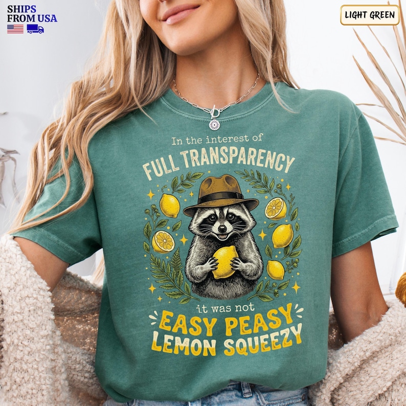 May include: A light green t-shirt featuring a graphic of a raccoon wearing a brown fedora hat and holding a yellow lemon. The text reads In the interest of FULL TRANSPARENCY it was not EASY PEASY LEMON SQUEEZY, surrounded by lemon illustrations.