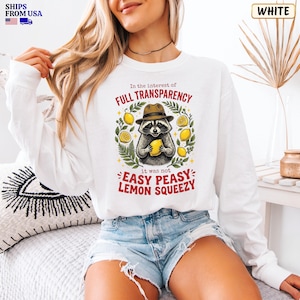 May include: A white long-sleeve sweatshirt featuring a graphic of a raccoon wearing a brown hat and holding a yellow lemon. The text reads In the interest of FULL TRANSPARENCY it was not EASY PEASY LEMON SQUEEZY, surrounded by green leaves.