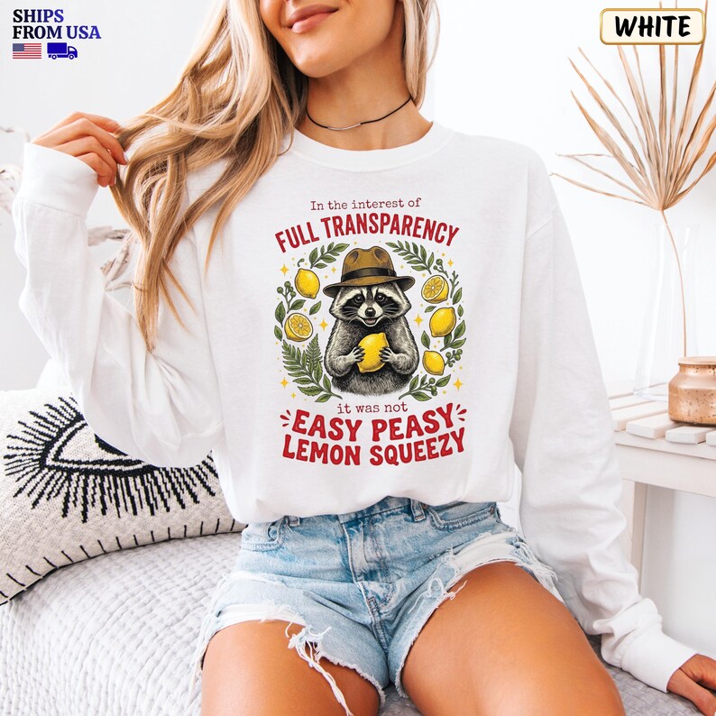 May include: A white long-sleeve sweatshirt featuring a graphic of a raccoon wearing a brown hat and holding a yellow lemon. The text reads In the interest of FULL TRANSPARENCY it was not EASY PEASY LEMON SQUEEZY, surrounded by green leaves.