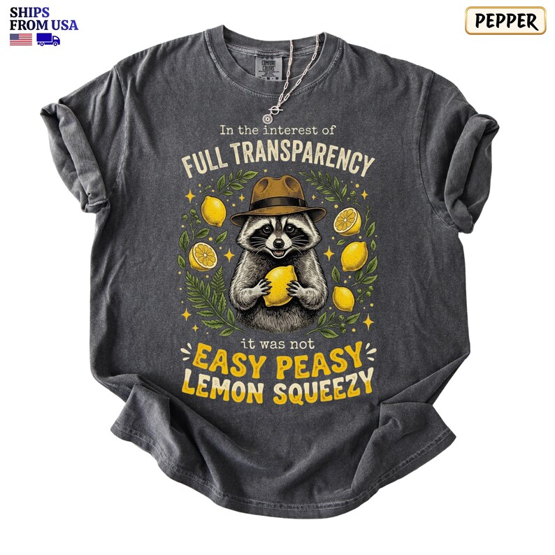 May include: A charcoal gray t-shirt featuring a graphic of a raccoon wearing a brown hat and holding a yellow lemon. The text reads In the interest of FULL TRANSPARENCY it was not EASY PEASY LEMON SQUEEZY, surrounded by yellow lemons and green leaves.
