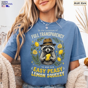 May include: A blue denim-colored t-shirt featuring a graphic of a raccoon wearing a brown fedora hat. The raccoon holds a yellow lemon, surrounded by lemon slices and green leaves. Text reads: In the interest of FULL TRANSPARENCY it was not EASY PEASY LEMON SQUEEZY.