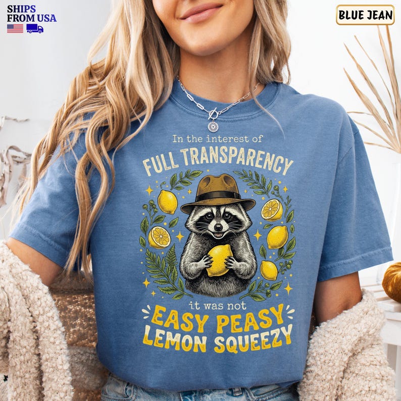 May include: A blue denim-colored t-shirt featuring a graphic of a raccoon wearing a brown fedora hat. The raccoon holds a yellow lemon, surrounded by lemon slices and green leaves. Text reads: In the interest of FULL TRANSPARENCY it was not EASY PEASY LEMON SQUEEZY.