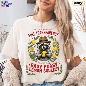 May include: An ivory t-shirt featuring a graphic of a raccoon wearing a brown hat and holding a yellow lemon. The text reads: In the interest of FULL TRANSPARENCY it was not EASY PEASY LEMON SQUEEZY. The design includes green leaves and yellow lemons.