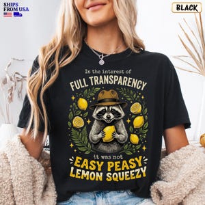 May include: A black t-shirt featuring a graphic of a raccoon wearing a brown fedora hat and holding a yellow lemon. The text reads In the interest of FULL TRANSPARENCY it was not EASY PEASY LEMON SQUEEZY, surrounded by yellow lemons and green leaves.