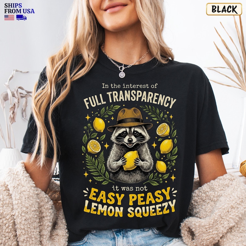 May include: A black t-shirt featuring a graphic of a raccoon wearing a brown fedora hat and holding a yellow lemon. The text reads In the interest of FULL TRANSPARENCY it was not EASY PEASY LEMON SQUEEZY, surrounded by yellow lemons and green leaves.