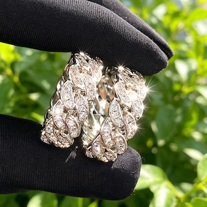 May include: Two silver rings with a linked chain design, encrusted with numerous small, sparkling clear stones. The rings are held against a black background with a blurred green foliage backdrop, creating a contrast that highlights the jewelry's brilliance.