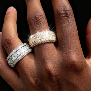 May include: Two rings, one gold and one silver, are displayed on a hand. The gold ring is encrusted with small, sparkling stones and has a detailed design. The silver ring has a similar design, with a braided pattern of stones.