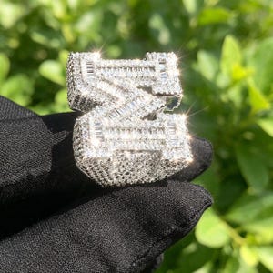 Könnte beinhalten: Ein funkelnder, silberfarbener Ring in Form des Buchstabens "Z", besetzt mit zahlreichen kleinen, klaren Edelsteinen. Der Ring wird vor einem schwarzen Hintergrund mit verschwommenem Grün gehalten. Das Design kombiniert Baguette- und Rundschliffe.
