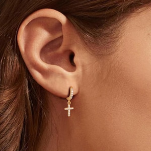 May include: A gold-colored hoop earring with small, clear stones and a dangling cross pendant. The cross and the hoop are encrusted with small, clear stones. The earring is worn on a person's ear.