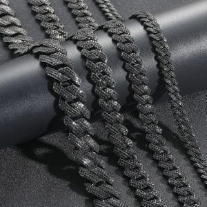 May include: Close-up of several black, chunky link chains. The chains are made of small, square-cut black stones, giving them a sparkling appearance. The chains are arranged on a dark surface, creating a contrast that highlights their design.