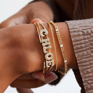 May include: A collection of gold bracelets adorns a wrist. One bracelet spells out "CHLOE" in sparkling crystals. Other bracelets include a delicate chain and a curb chain with small, clear stones. The bracelets are layered and create a stylish look.