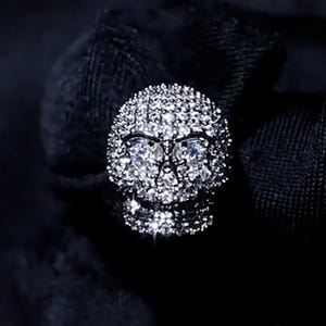 May include: A skull-shaped ring covered in small, sparkling, clear gemstones. The ring has a dark background, and the skull's eye sockets are visible. The skull's features are well-defined, and the gemstones catch the light.