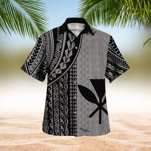 May include: Short-sleeved button-down shirt with a black and gray tribal pattern. The shirt features a black collar and a central design with a black symbol. The shirt is displayed on a sandy beach background.