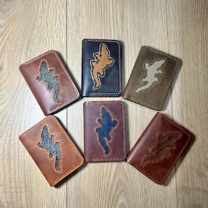 May include: A collection of six leather wallets in various shades of brown, each featuring a lizard-shaped appliqué in contrasting colors. The wallets are arranged on a light wooden surface, showcasing the different color combinations and textures.