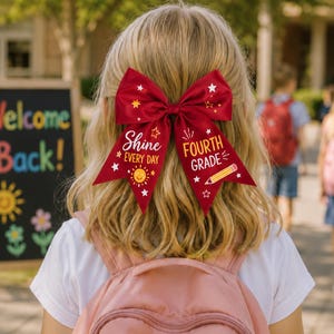 Könnte beinhalten: Eine große rote Haarschleife mit weißen und gelben Grafiken von Sternen, einer Sonne und einem Bleistift. Der Text auf der linken Seite lautet Shine EVERY DAY und auf der rechten Seite FOURTH GRADE. Die Schleife besteht aus glattem Satin-Stoff.