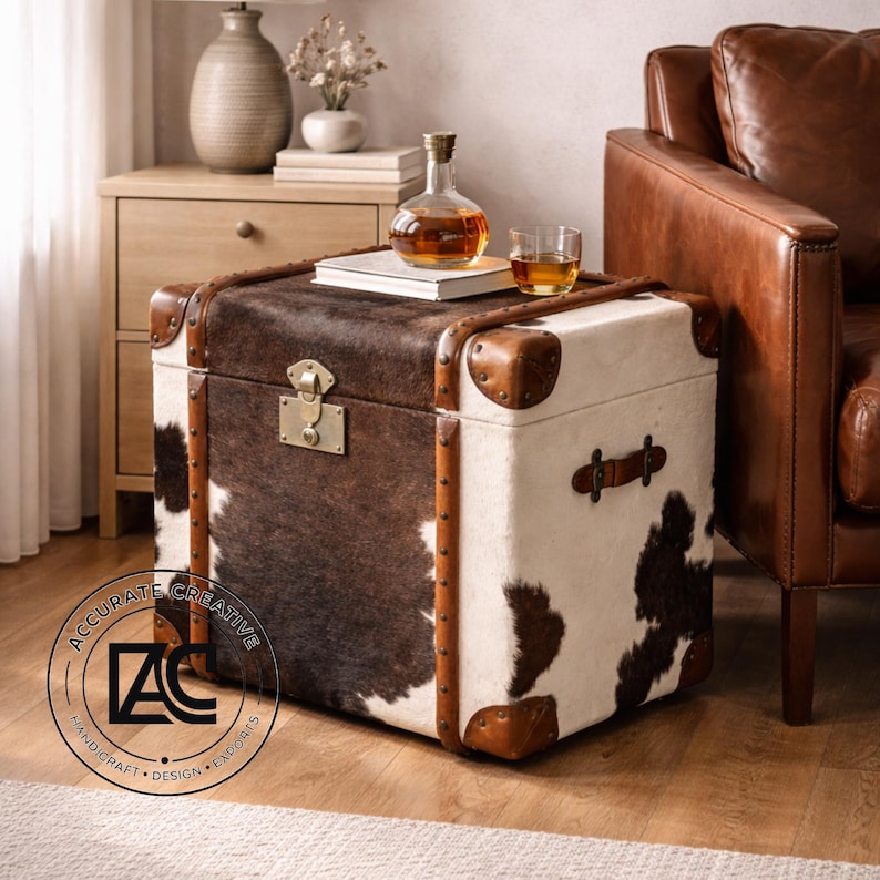 May include: A decorative trunk with a cowhide pattern, brown leather trim, and brass hardware. The trunk is used as a side table, with a bottle of liquor and a glass on top. The trunk is next to a brown leather armchair.