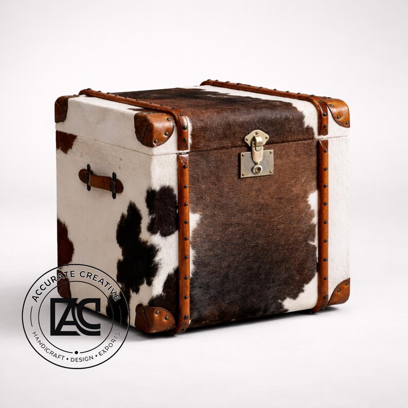 May include: A square trunk with a cowhide exterior, featuring a mix of white, brown, and black patches. The trunk has brown leather trim, corner protectors, and a metal clasp. A leather handle is visible on the side.