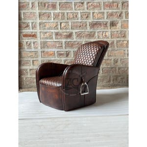 May include: A brown leather armchair featuring a diamond-quilted pattern on the backrest and seat. The chair includes a silver-toned metal stirrup accent attached to the side panel. A circular logo with the text ZC is visible on the side.