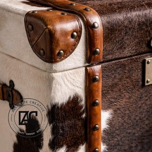 May include: Close-up of a decorative trunk with a cowhide exterior. The trunk features brown and white fur, accented with brown leather trim and brass studs. The corners are reinforced with leather and metal hardware.