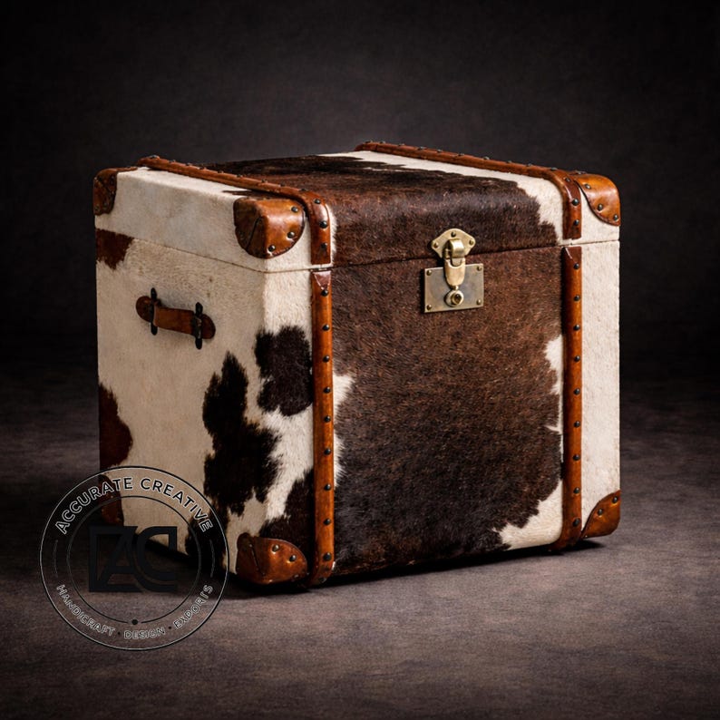 May include: A square trunk with a cowhide exterior, featuring brown and white patches. The trunk has brown leather trim, metal corner protectors, and a brass lock. A leather handle is visible on the side. The overall design is rustic and vintage.