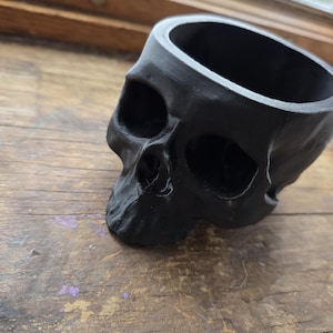 May include: A black skull-shaped planter made of a solid material. The planter has a hollow interior and is designed with detailed facial features, including eye sockets, a nose cavity, and a jawline. The planter is sitting on a wooden surface.