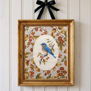Op de afbeelding: Een rechthoekige gouden lijst met een stoffen paneel met een centrale ovale patch van een blauwe vogel op een tak. De achtergrondstof heeft een patroon van herfstbladeren en bessen. Aan de bovenkant hangt een zwarte strik van lint.