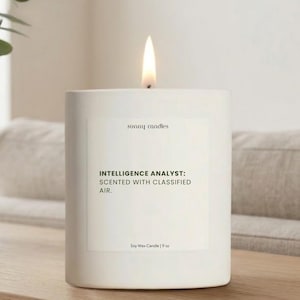 May include: A white soy wax candle with a lit flame. The candle has a rectangular label with the text "INTELLIGENCE ANALYST: SCENTED WITH CLASSIFIED AIR." The label also includes the text "sunny candles" and "Soy Wax Candle | 9 oz."