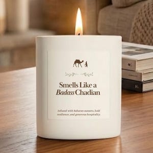 May include: A white cylindrical candle with a lit wick. The label features a brown camel icon, a green leaf graphic, and the text: Smells Like a Badass Chadian. Below this, it reads: Infused with Saharan sunsets, bold resilience, and generous hospitality.