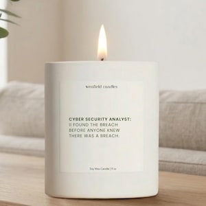 May include: A white soy wax candle with a lit flame. The candle has a rectangular label with the text "CYBER SECURITY ANALYST: I FOUND THE BREACH BEFORE ANYONE KNEW THERE WAS A BREACH." The candle is 9 oz.