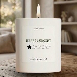 May include: A lit white candle with the text "wenfield candles" and "HEART SURGERY" on a label. The label includes a one-star rating and the words "Do not recommend". The candle is on a wooden surface.