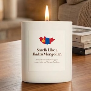 May include: A white cylindrical candle with a lit wick. The label features a red, blue, and yellow map of Mongolia, the text "Smells Like a Badass Mongolian," and the phrase "Infused with endless steppes, horse trails, and fearless freedom."